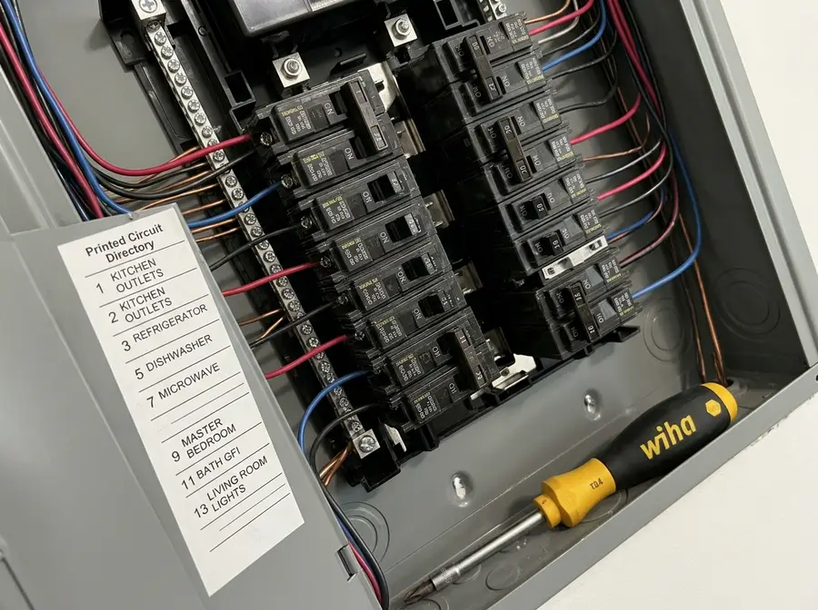 Tight diagonal crop of a finished 200-amp panel interior with two rows of labelled breakers, a printed circuit directory and a torque screwdriver resting on the panel lip, everything installed and connected